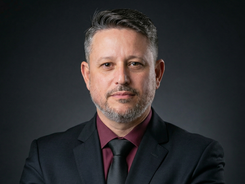 Retrato corporativo de sócio-diretor da Fenicio, utilizando terno preto, gravata preta e camisa vinho, com olhar focado e postura de autoridade sobre fundo escuro.