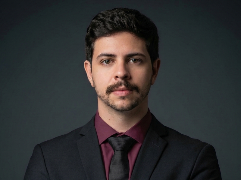 Retrato corporativo de especialista da Fenicio, vestindo terno preto, gravata slim preta e camisa vinho, com expressão séria e profissional sobre fundo neutro.