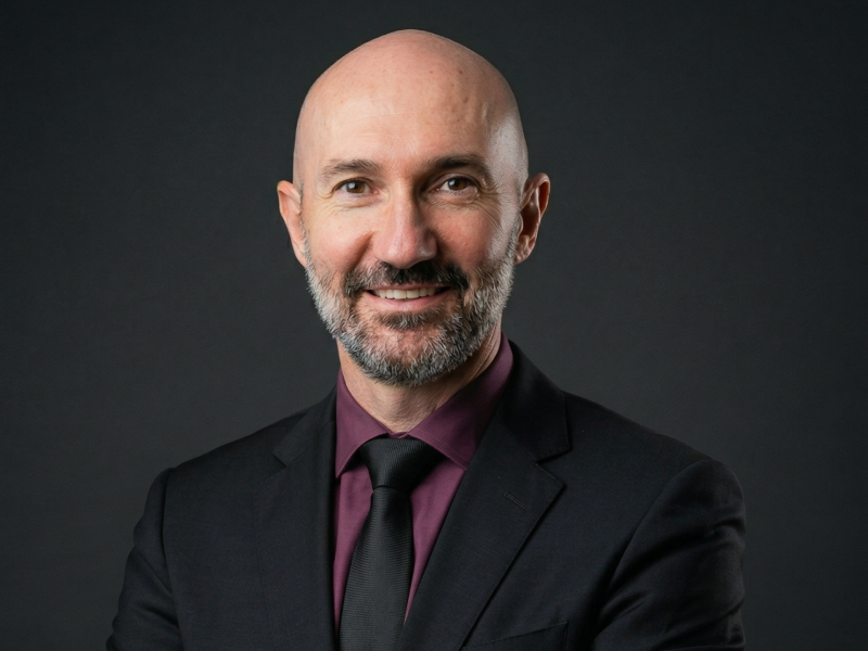 Retrato corporativo do sócio-fundador da Fenicio, vestindo terno preto, gravata preta e camisa vinho, sorrindo sobre fundo cinza escuro.