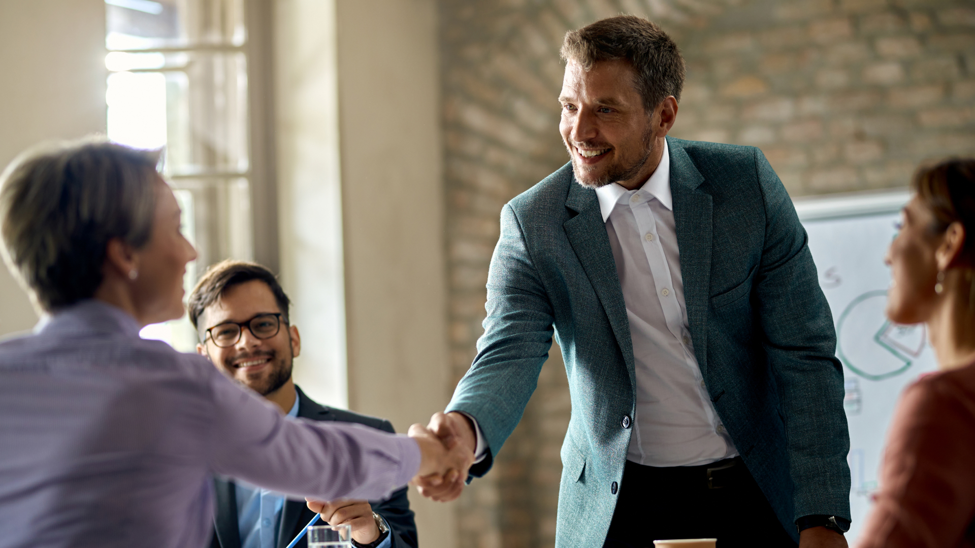 Especialista da Fenicio apertando a mão de uma profissional após workshop corporativo, com equipe sorrindo ao fundo em ambiente de escritório.