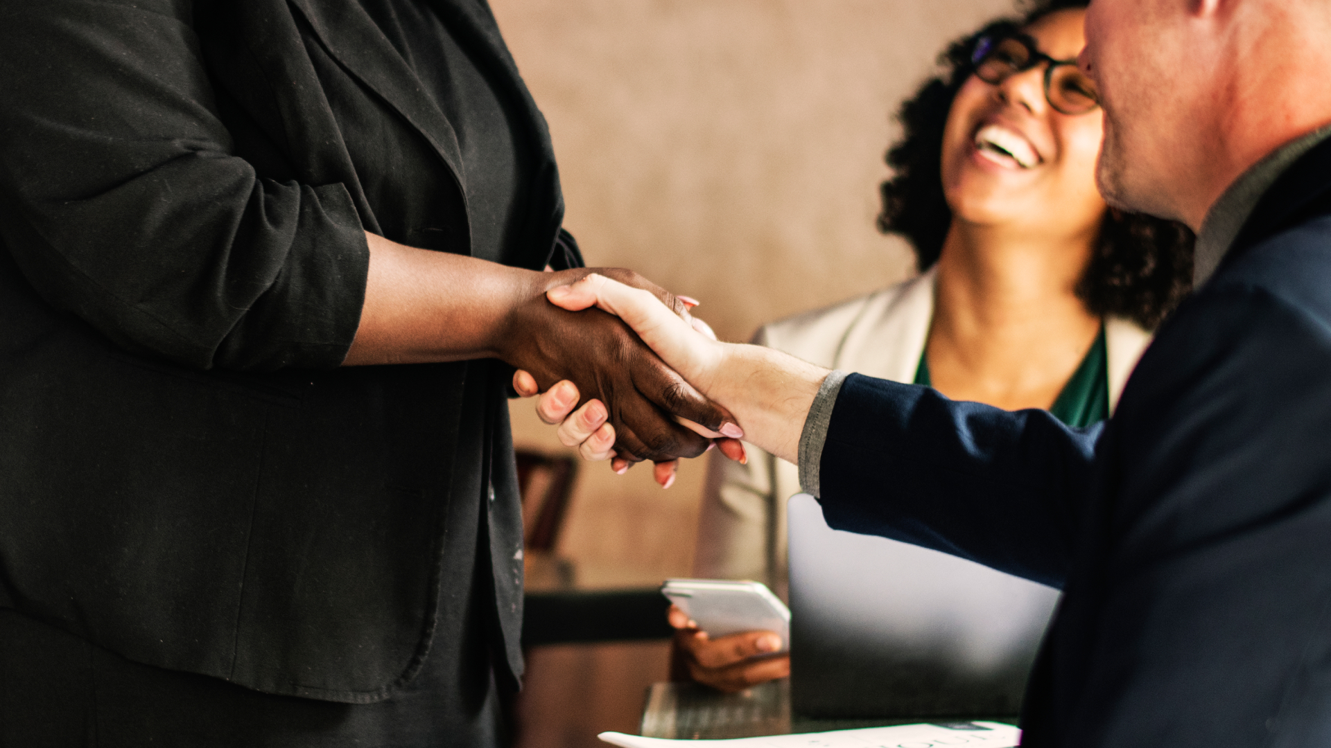 Aperto de mão firme entre consultor e cliente após conclusão de treinamento executivo da Fenicio, com profissional sorrindo ao fundo simbolizando satisfação.