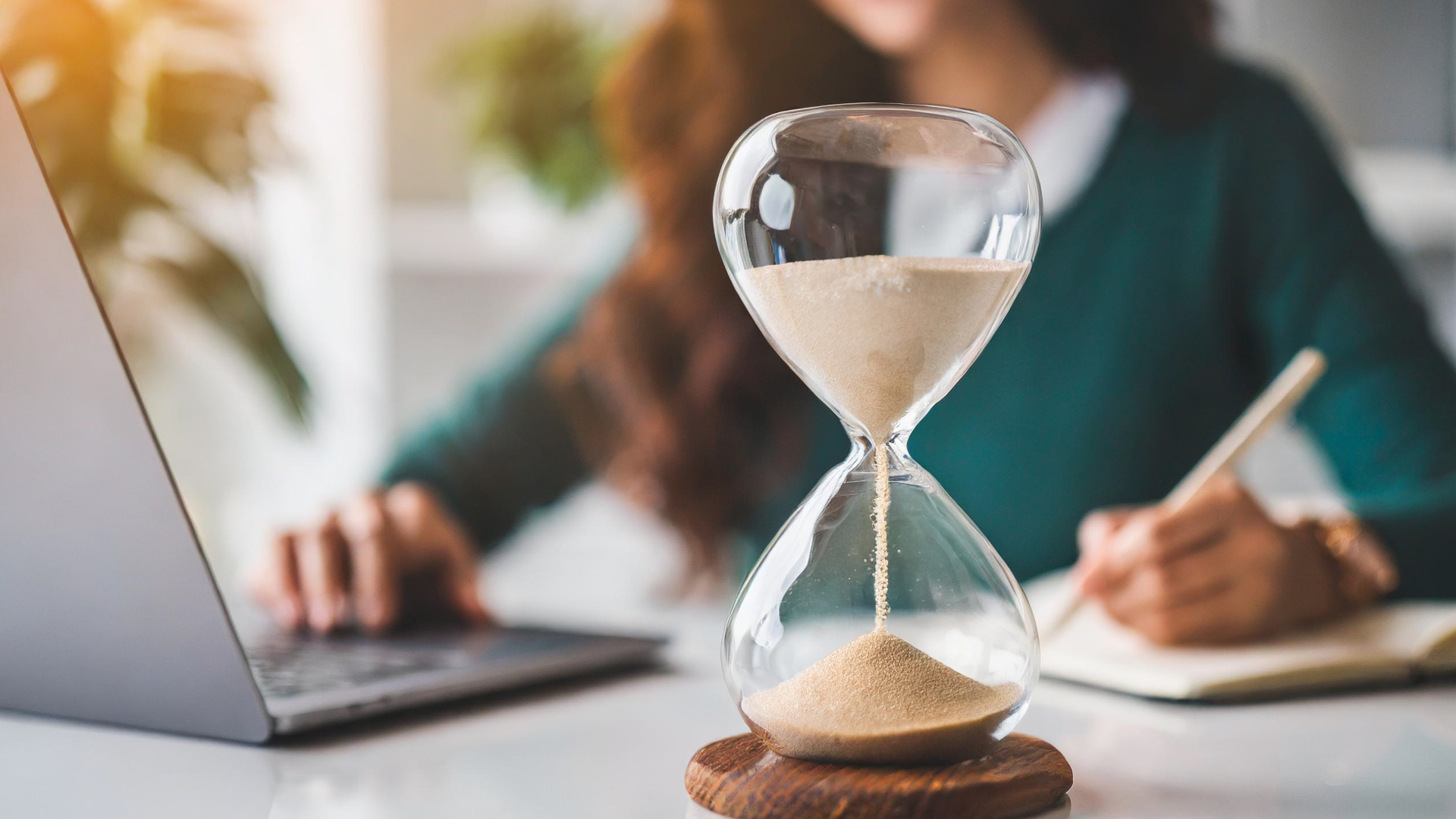 Ampulheta de vidro em primeiro plano com profissional trabalhando ao fundo, simbolizando a gestão de tempo e produtividade nos treinamentos da Fenicio.