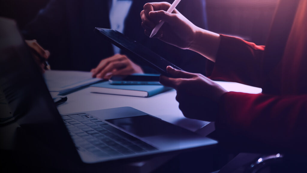 Duas pessoas em uma reunião de negócios analisando dados em um tablet e notebook, com iluminação dramática em tons de azul e roxo, simbolizando consultoria estratégica.